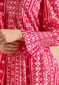 Różowa sukienka w wzory z cechami kwiatowymi, muszelkowymi i geometrycznymi. Długie rękawy z plisowanymi detalami, miękka tkanina z płynącą sylwetką.