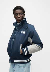 Άνδρας με μπλε σκούρο και γκρι μπουφάν North Face με ριγέ μανσέτες και ανοιχτό μπλε τζιν παντελόνι, στέκεται με το χέρι στην τσέπη, κοιτάζοντας προς τα πλάγια.