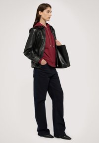 Černá kožená bunda přes vínový mikinu, doplněná tmavě modrými džíny a černými špičatými botami. Stojící pozice, drží černou tašku.