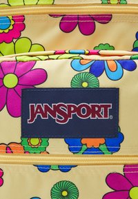Sárga hátizsák élénk virágmintákkal rózsaszín, kék, narancs és zöld színekben. Sötétkék JanSport logóval és sárga cipzárokkal rendelkezik.