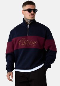 Sweatshirt bicolore marine et bordeaux, avec un col zippé mi-haut, un texte brodé en doré et des poignets côtelés. Texture douce, coupe oversize.