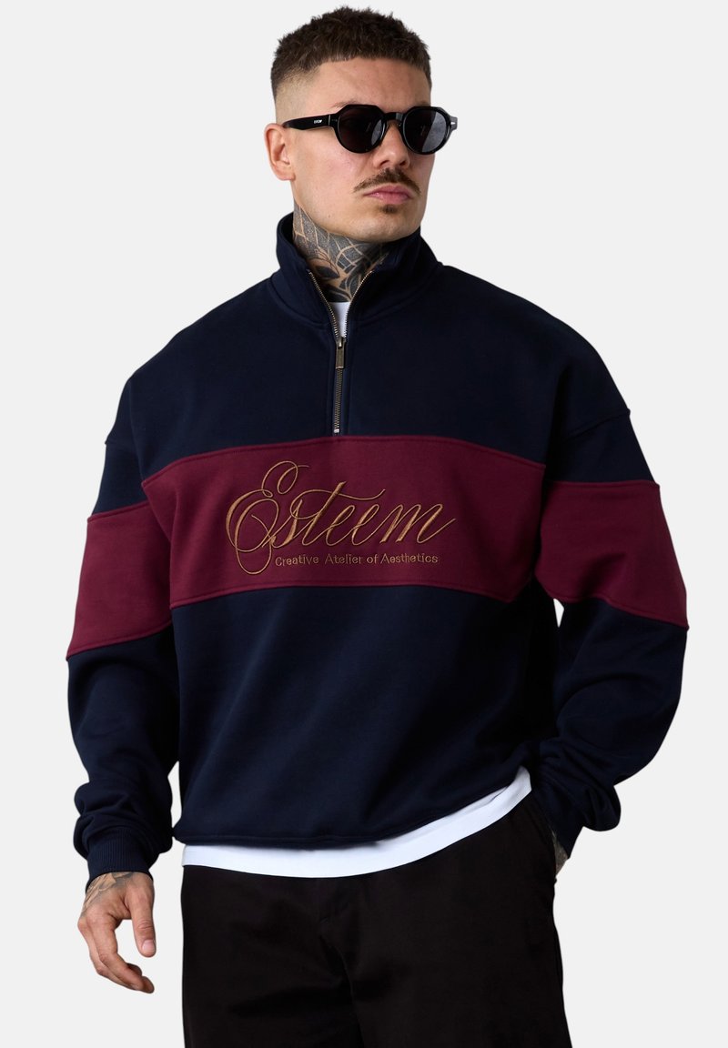 Sweatshirt bicolore marine et bordeaux, avec un col zippé mi-haut, un texte brodé en doré et des poignets côtelés. Texture douce, coupe oversize.