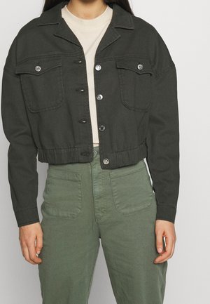 Nainen pukeutunut mustaan lyhyeen takkiin, jossa on nappikiinnitteiset taskut, beigen paidan päällä, yhdistettynä korkeavyötäröisiin vihreisiin housuihin, joissa on etutaskut.