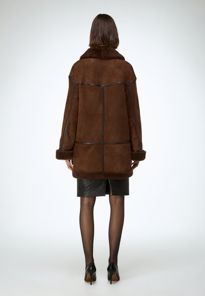 Bruine suède jas met een bontkraag, voorzien van zwarte leren biezen en lange mouwen. Gedragen over een zwarte leren rok, staand op hoge hakken.