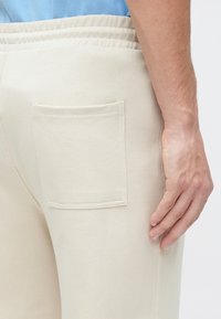 Pantalones de chándal beige con un solo bolsillo tipo parche en el lado derecho, cintura elástica y textura de tela suave.