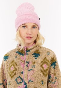 Blonde woman wearing a pink knit beanie and a beige fleece jacket with green, pink, and blue geometric patterns, looking at the camera.