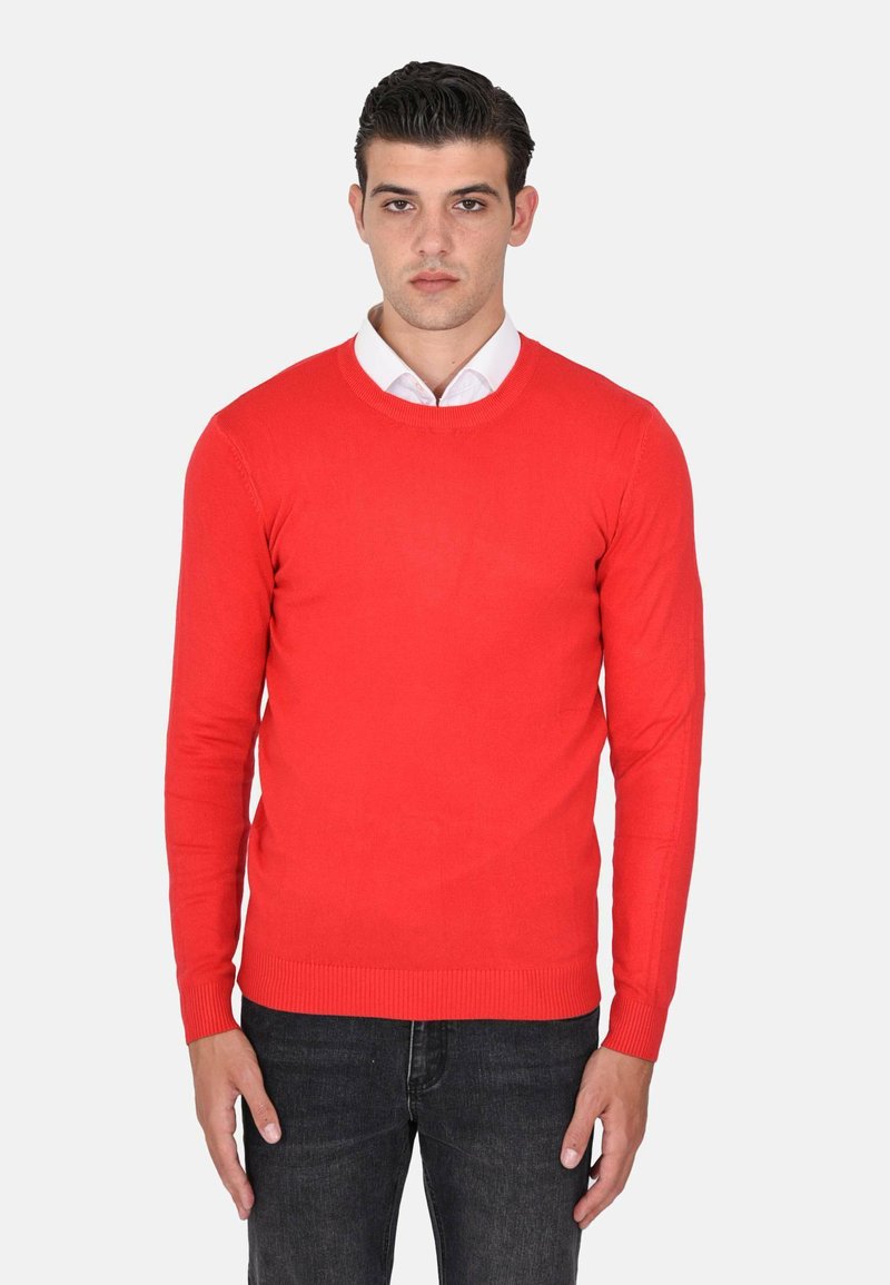 Maglione rosso lavorato a maglia con maniche lunghe, scollo rotondo e polsini e orlo a coste. Indossato sopra una camicia bianca con colletto, abbinato a jeans scuri.