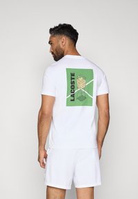 Λευκό βαμβακερό μπλουζάκι με ένα μεγάλο πράσινο σχέδιο στην πλάτη που απεικονίζει μια τσάντα με μπάλες τένις και το κείμενο "LACOSTE". Κοντά μανίκια, χαλαρή εφαρμογή.