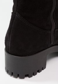 Bota de tobillo de ante negro con suela de goma gruesa. Cuenta con un cierre lateral y un upper texturizado con sutiles detalles de costura, diseñado para la durabilidad.