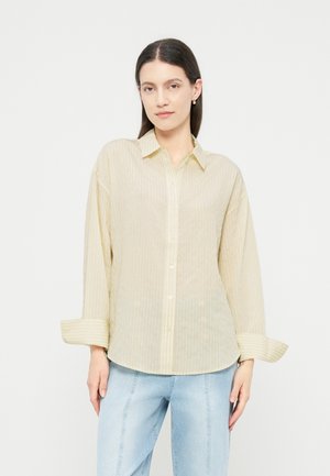Kvinde med langt mørkt hår bærer en løs, beige stribet knap-op-skjorte med op-rullede manchetter og lyseblå jeans mod en hvid baggrund.