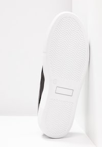 Semelle d'une basket noire et blanche avec un motif de crêpe en chevrons, taille 36 indiquée sur la semelle, appuyée contre un mur blanc.