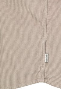 Beige Cord-Stoff mit vertikalen Rippungen, sichtbarer Naht und einem kleinen weißen Etikett mit der Aufschrift "EIGHT2NINE", das in der Nähe der Kante genäht ist.