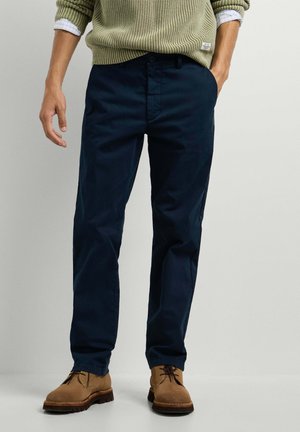 Uomo che indossa pantaloni blu scuro a taglio dritto, scarpe marroni con lacci, maglione verde oliva sopra una camicia bianca, in piedi con una mano in tasca.