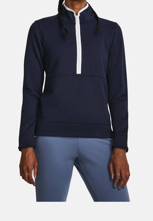 Personne portant un pull à demi-fermeture éclair bleu marine avec une fermeture éclair blanche et un pantalon bleu clair ajusté, debout les bras détendus le long du corps.