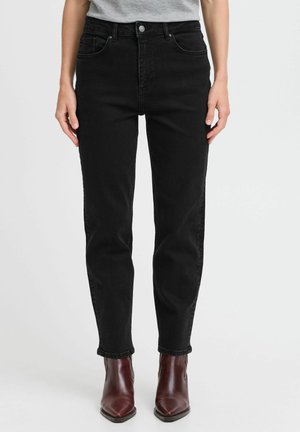 Person iført højtaljede sorte jeans og brune spidse ankelstøvler, stående foran en ensfarvet lys grå baggrund.