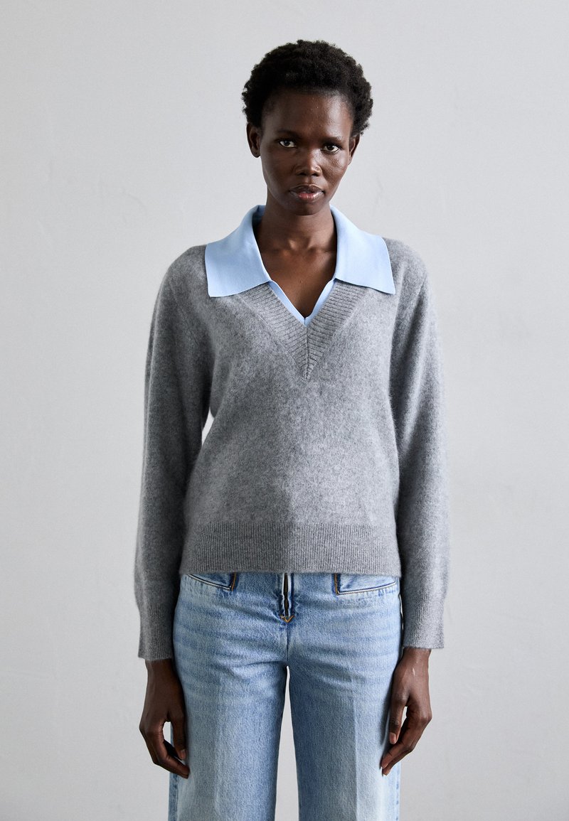 Suéter de lana gris con un cuello de camisa azul claro, diseño de escote en V, mangas largas y un sutil detalle de canalé en el dobladillo. Combinado con jeans azul claro.
