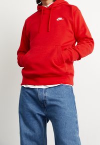 Röd huvtröja i mjukt tyg, med en framficka, dragsko på huven och vit logotyp på bröstet. Matchas med blå denimjeans.