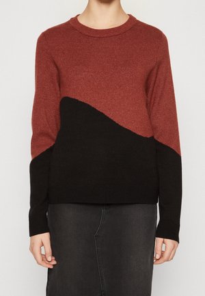 Femme portant un pull en tricot bicolore rouille et noir à manches longues et col rond, associé à une jupe foncée.