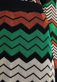 Stickad tyg med zigzag-mönster i svart, rostorange, grön och beige färger, som visar detaljerad textur och stygnarbete.