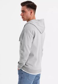 Haut à capuche gris clair avec une poche frontale, un capuchon à cordon et des poignets côtelés. Texture lisse et design décontracté qui se marie bien avec un jean bleu.