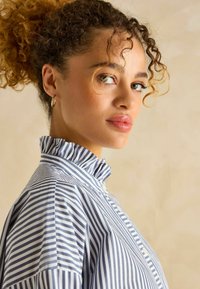 Sinine ja valge triibuline pluus, millel on volangiga krae ja nööpide sulgemine. Pehme tekstuur, regulaarne lõige ja pikad varrukad.