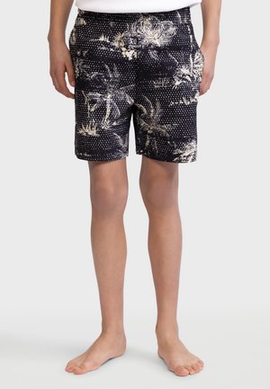 Homme portant un short noir avec des motifs de palmiers tropicaux beiges et des points, debout pieds nus devant un fond clair uni.