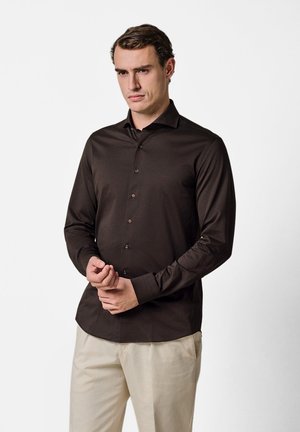 Mand iført en tætsiddende mørkebrun langærmet skjorte med knapper og lyse beige bukser, der justerer sit venstre håndled med sin højre hånd.