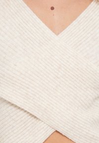 Roomkleurige omslagtrui met een geribbelde textuur, voorzien van een V-hals en een diagonale overlap.