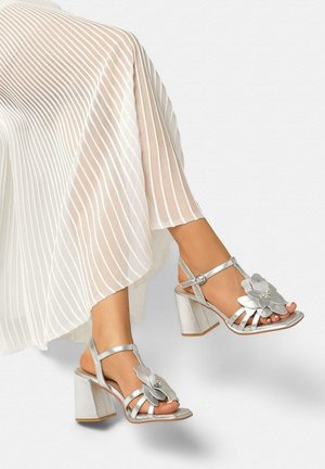 Pies con sandalias de tacón plateadas con decoraciones de flores, combinadas con una falda blanca plisada.