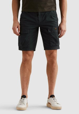 Man wearing black cargo shorts with side pockets and white sneakers, standing against a plain gray background.