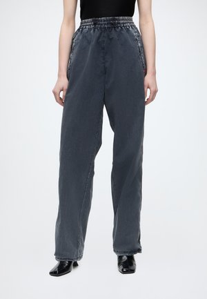 Personne portant un pantalon ample gris foncé taille haute avec une ceinture élastique et des chaussures à talons noires, debout devant un fond uni.