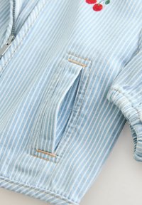Helleblaue gestreifte Denimjacke mit einem Reißverschluss, die ein kleines gesticktes Kirschdesign und eine seitliche Tasche aufweist.