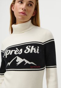 Jeune femme portant un pull à col roulé blanc et noir avec "Après Ski" et des motifs de montagne sur la poitrine.