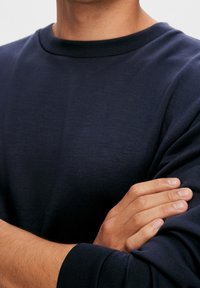 Mörkblå bomulls sweatshirt med rund nacke, långa ärmar och en slät textur. Tröjan bärs med armarna korsade.