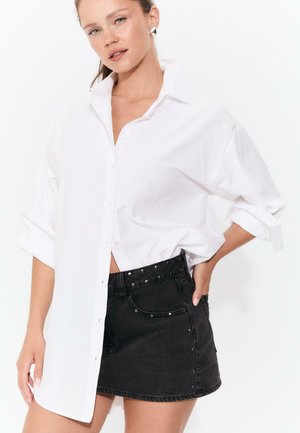 Donna che indossa una camicia bianca oversize con maniche arrotolate e una minigonna di jeans nera con borchie, posando con una mano sulla vita.