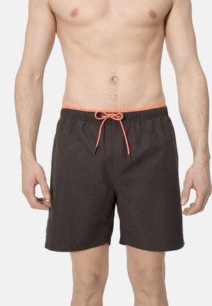 Hombre con pantalones cortos de baño negros con cordón naranja, de pie contra un fondo claro y liso, torso visible desde el pecho hasta las rodillas.