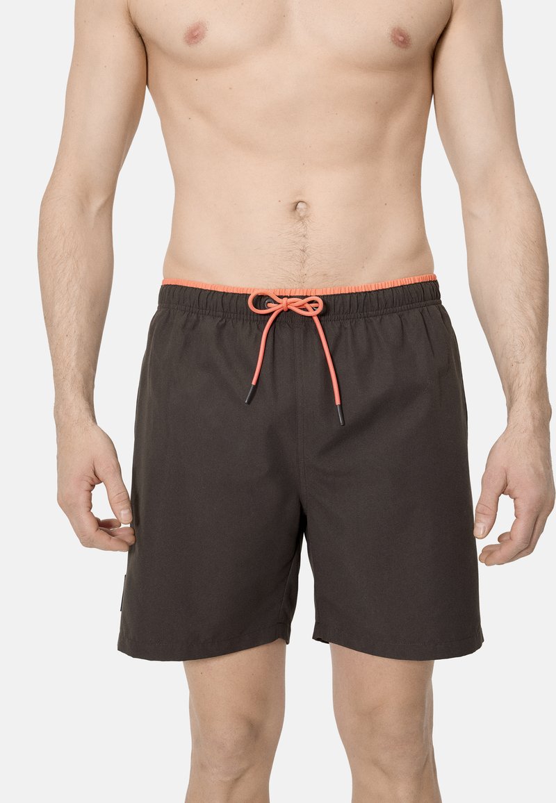 Hombre con pantalones cortos de baño negros con cordón naranja, de pie contra un fondo claro y liso, torso visible desde el pecho hasta las rodillas.
