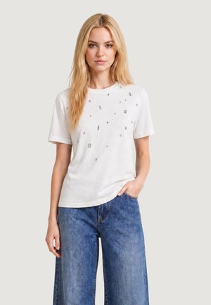 Blond kvinde iført en hvid kortærmet skjorte med små spredte pynt og løse blå denimjeans, stående med den ene hånd i lommen.