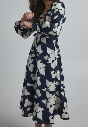 Žena oblečená v tmavě modrých midi šatech s bílým květinovým potiskem, s dlouhými rukávy a zavázáním v pase, pózující před neutrálním šedým pozadím.
