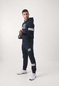 Námornícky súprava mikiny a nohavíc s bielymi pruhmi, potlačou loga a manžetami s rebrovaním. Model drží čierny kettlebell a má na sebe biele tenisky.