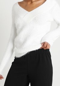 Weißer, flauschiger Pullover mit V-Ausschnitt und langen Ärmeln, kombiniert mit schwarzen, strukturierten Hosen mit elastischem Bund.