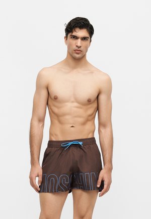 Fiatal férfi sötét hajjal, barna fürdőnadrágot visel kék megkötővel és „Moschino” felirattal, egyszerű világos háttér előtt.