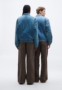 Dvaja ľudia sedia chrbtom, oblečení v svetlomodrých denimových bundách s elastickými lemami a širokými hnedými nohavicami. Bundy majú voľný strih a minimálne ozdoby.