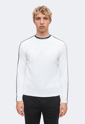 Jonge man met blond haar die een wit shirt met lange mouwen met zwarte bies en zwarte broek draagt, staand tegen een effen lichte achtergrond.