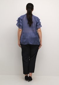 Světle modrá plisovaná blůza s volánovými rukávy, kombinovaná s černými tapered kalhotami a černými vysokými podpatky. Minimalistický design.