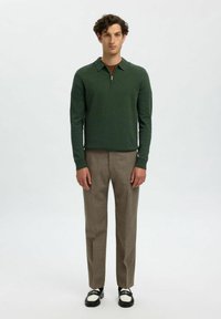 Grøn lynlåspullover med lange ærmer, parret med beige bukser. Fodtøjet har et design i sort og hvidt to-tone. Enkelt, moderne pasform.