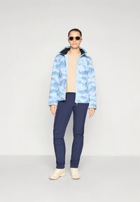 Mėlynos ryškios vandeniui atsparios striukės, užsegama užtrauktuku, su dviem priekinėmis kišenėmis, derinama su smėlio spalvos palaidine ir tamsiai mėlynais kelnėmis, papildoma šviesios spalvos avalyne.