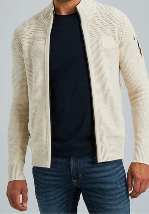 Hombre con cárdigan beige texturizado con cremallera sobre camisa negra y jeans azules, de pie frente a un fondo liso.