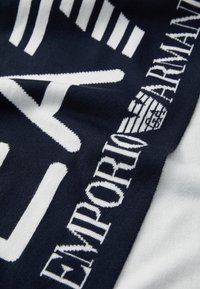 Eșarfă bleumarin cu branding alb contrastant. Materialul pare moale și tricotat, având o finisare texturată și detalii vizibile ale logo-ului.