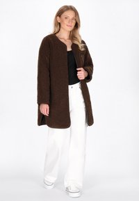 Cappotto marrone, morbido e oversize con colletto rotondo e zipper frontale, abbinato a pantaloni bianchi a gamba larga e sneaker casual.
